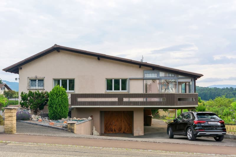 2-story house with stucco exterior, brown roof, large windows, wooden garage door, balcony, and a parked car in the driveway. The house is surrounded by landscaping with shrubs and trees.
