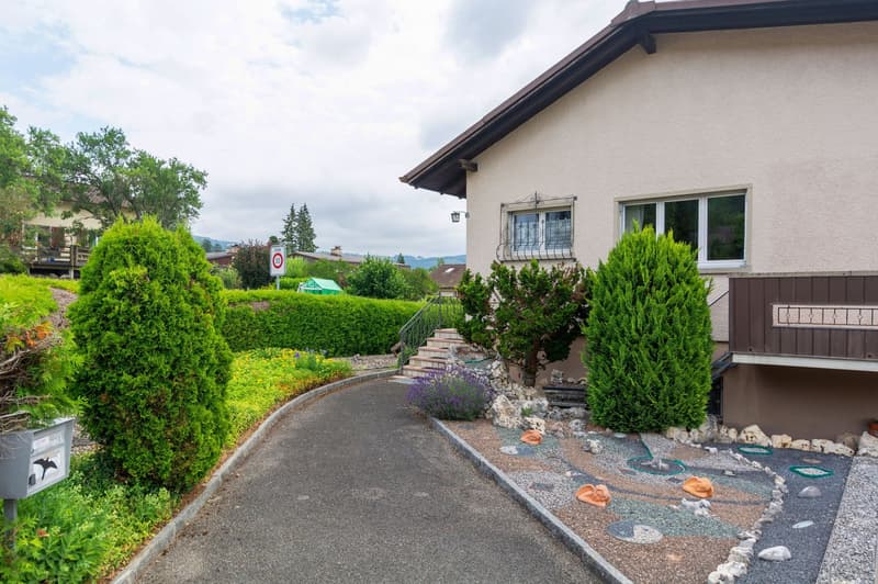 Two-story house with stucco exterior, brown roof, and large windows. The house is surrounded by a well-maintained garden with lush greenery, including shrubs and trees. There is a paved walkway leading to the house, and a designated parking area is visibl