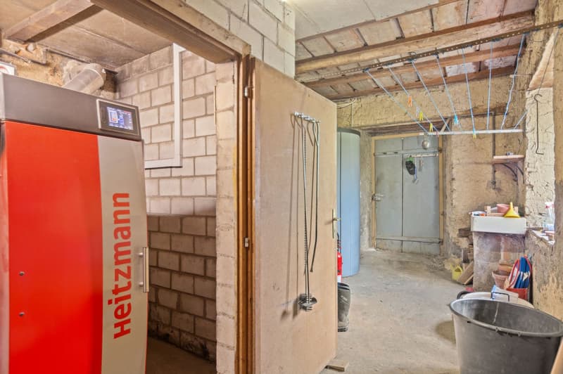 Unfinished basement or storage area with exposed brick walls, wooden beams, and a large red appliance, likely a washing machine or dryer. The space also contains various tools and equipment, suggesting it is used for storage or utility purposes.