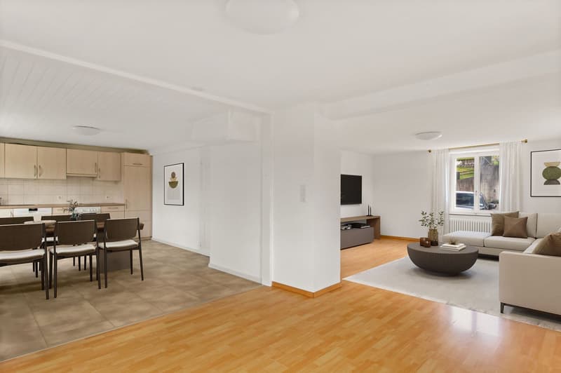 The image shows an open floor plan with a living room, dining area, and kitchen. The living room has a sofa, coffee table, and wall decor. The dining area has a table and chairs. The kitchen has light-colored cabinets and a dishwasher. The floors are a mi