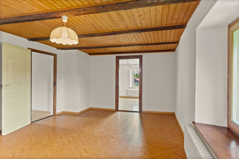 This image shows a spacious room with a wooden ceiling and exposed beams. The room has hardwood floors and a large window that provides natural light. There is a decorative light fixture hanging from the ceiling. The room appears to be empty, with no furn