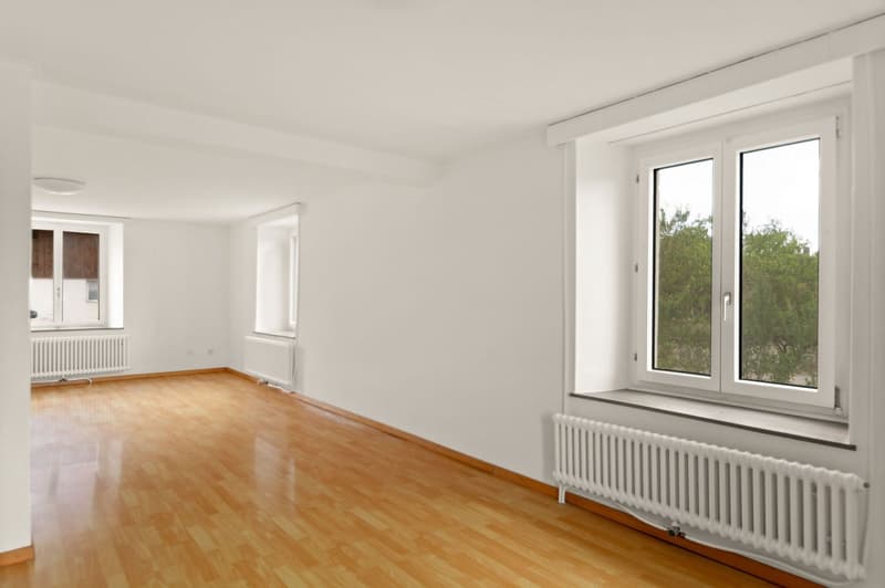 Spacious living room with hardwood floors, large windows providing a view of the surrounding greenery, and white walls. The room has a clean and minimalist design.
