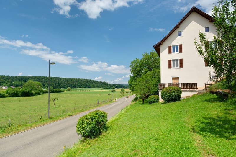 2-story residential building with white walls and brown roof, surrounded by green trees and grassy fields, with a paved road and street lamp in the foreground