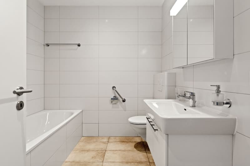 Bathroom: white tiled walls, white tiled floor, white bathtub, white sink, white toilet, mirror, soap dispenser, shower handle, white cabinets