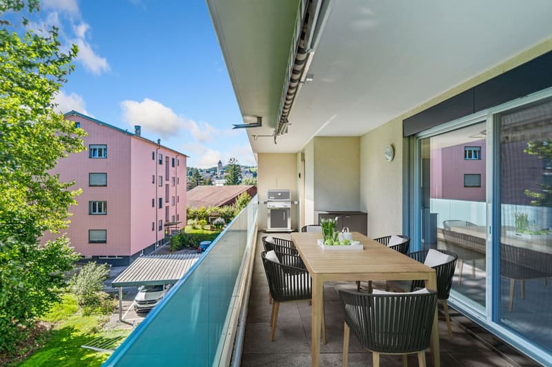 A balcony with a dining table and chairs, glass railings, glass door, and a view of the city and buildings.