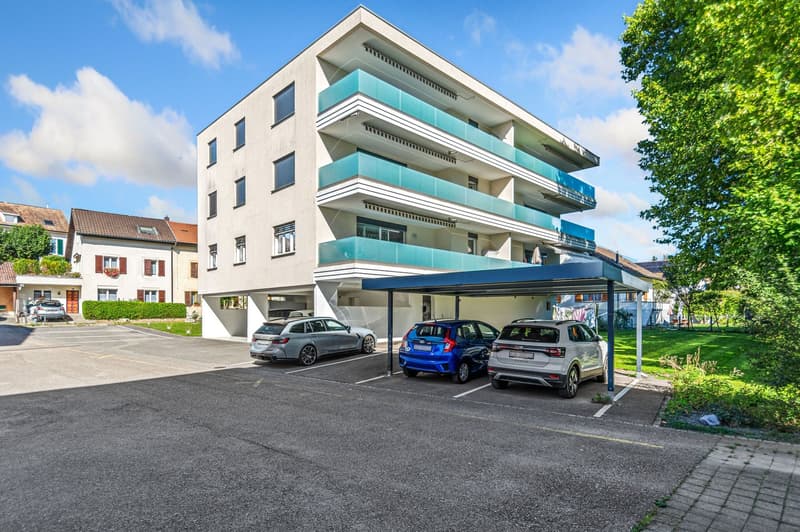 Modern apartment building with 4 stories, grey exterior, green balconies, car parking spaces, covered parking lot, greenery around.