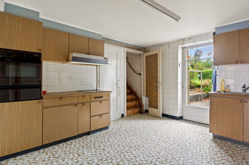 A kitchen with wooden cabinets, white tile walls, wooden staircase, large window, and black oven