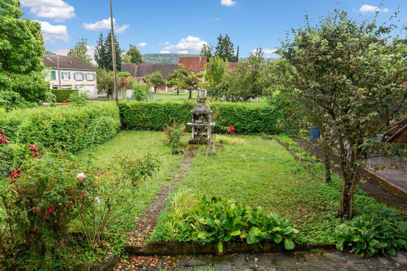 A picturesque garden with green grass, neatly trimmed hedges, a central water feature, vibrant flowers, and trees. The garden extends beyond the view to reveal a tranquil rural setting.