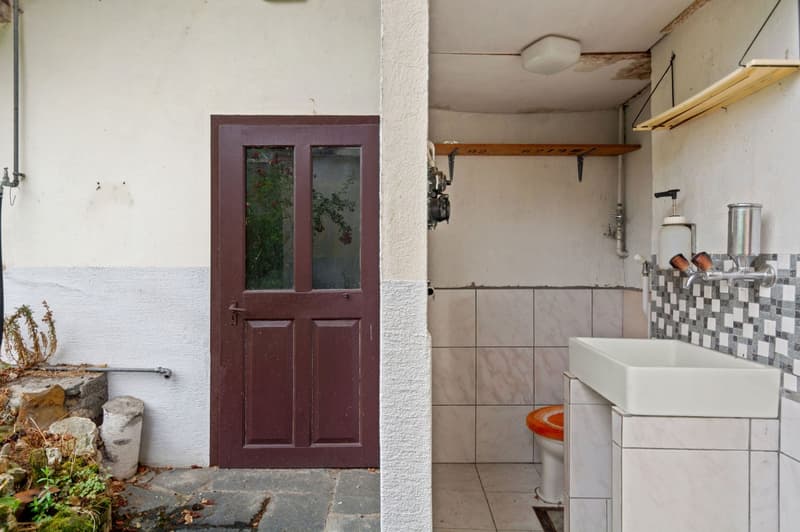 A bathroom with a sink, toilet, wall-mounted soap dispenser, and shelving. It has tiled walls and a door.