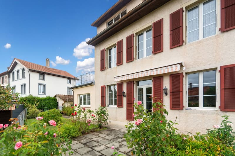 Exterior of a two-story house with red shutters and beige walls, surrounded by lush greenery and pink flowers.