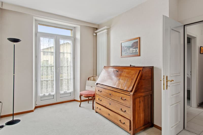 Empty room with white walls and beige flooring. Wooden desk with drawers. Glass doors leading to another room. Chair and picture frame on the wall.