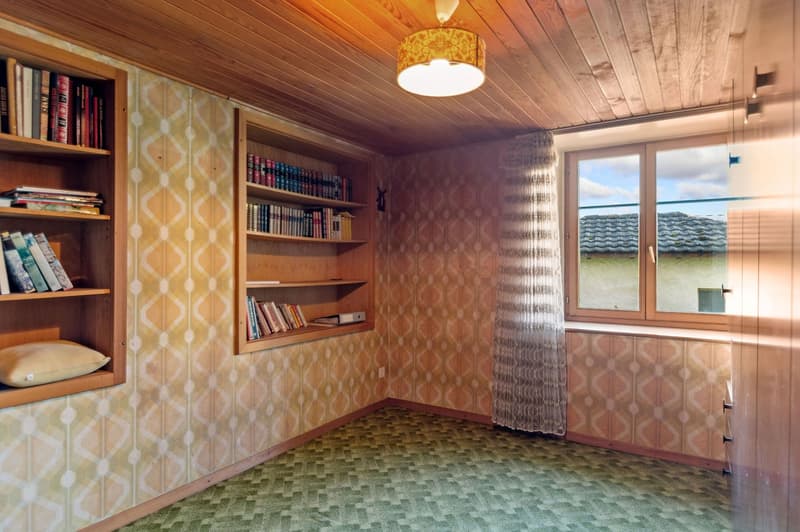 Empty room with wooden shelving, patterned wallpaper, carpet flooring, pendant light, and window with curtains.