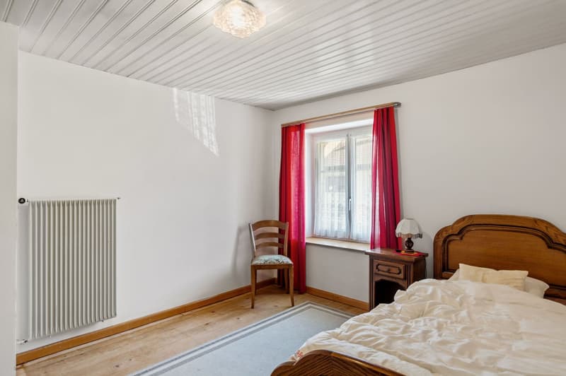 Wooden bed, white linens, wooden chair, nightstand with lamp, white radiator, wooden floor, red curtains