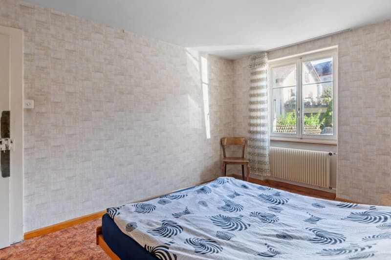 A bedroom with bed, bedspread, a wooden chair, a window with a curtain, and a radiator. The room has a white and beige wallpaper and wooden flooring.