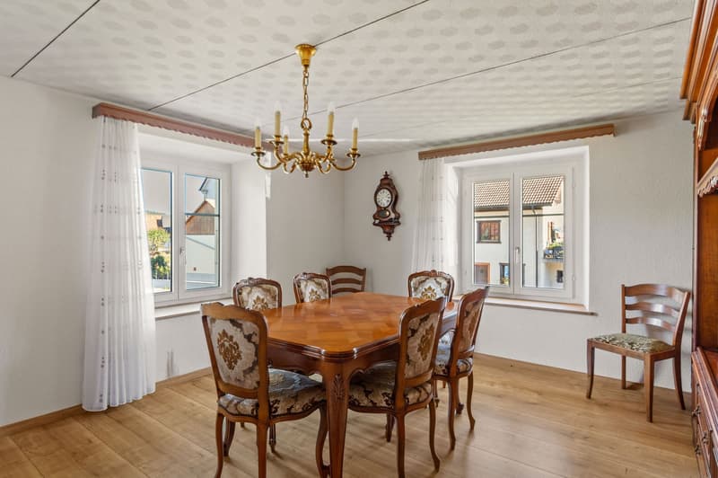 wooden dining table, chairs, chandelier, white walls, white curtains, clock, wooden cabinet