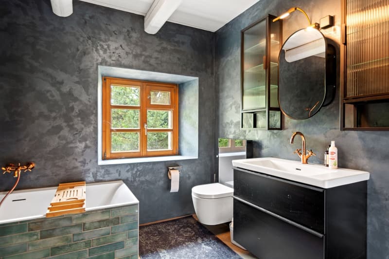 modern bathroom with grey wall, white bathtub with wooden tray, white toilet, grey floor tiles, white sink with wooden cabinets, mirrors, copper faucets, toilet roll, wooden window