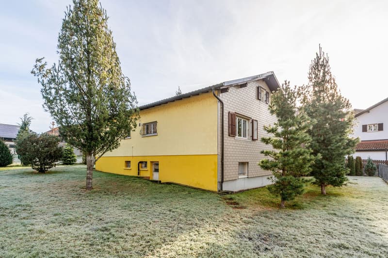 2 story house, yellow and white, windows, no visible entrance, fenced yard, pine trees