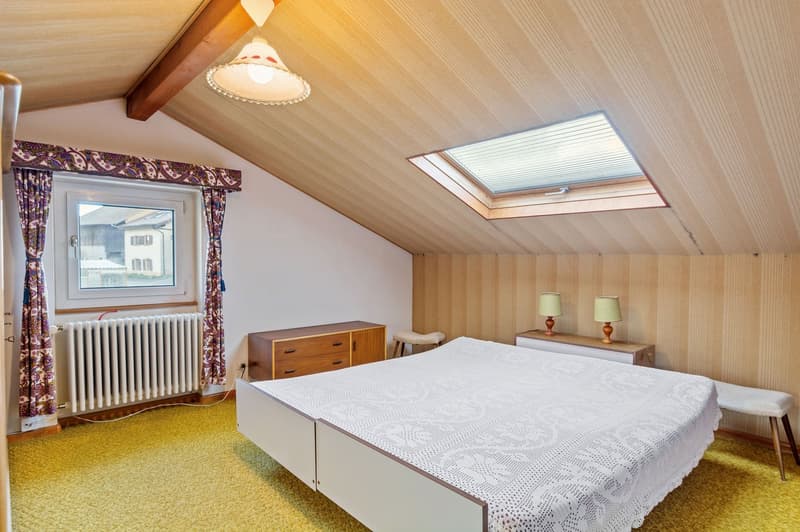 white bed, white crochet bedspread, white radiator, brown nightstands, lamps, white bench, wooden beams, yellow floor, small window, skylight