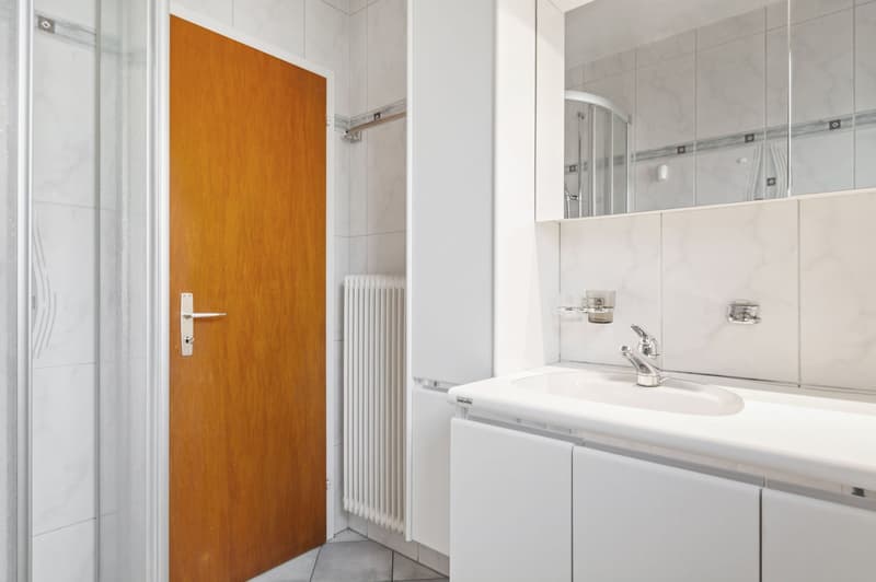 white walls, tiled floors, modern shower, toilet, sink with faucet, wooden door, white cabinets, silver mirror