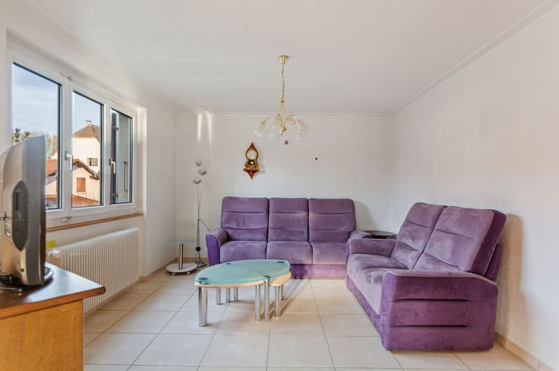Large room with white walls, tiled floor, two purple sofas, glass table, chandelier, radiator, television