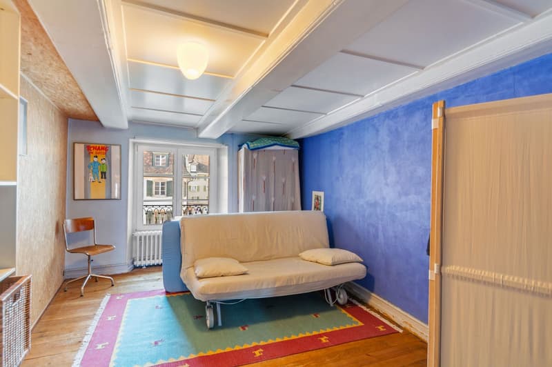 a cozy room with blue walls, a white couch with pillows, a wooden chair, a white radiator, a framed picture on the wall, and a carpet