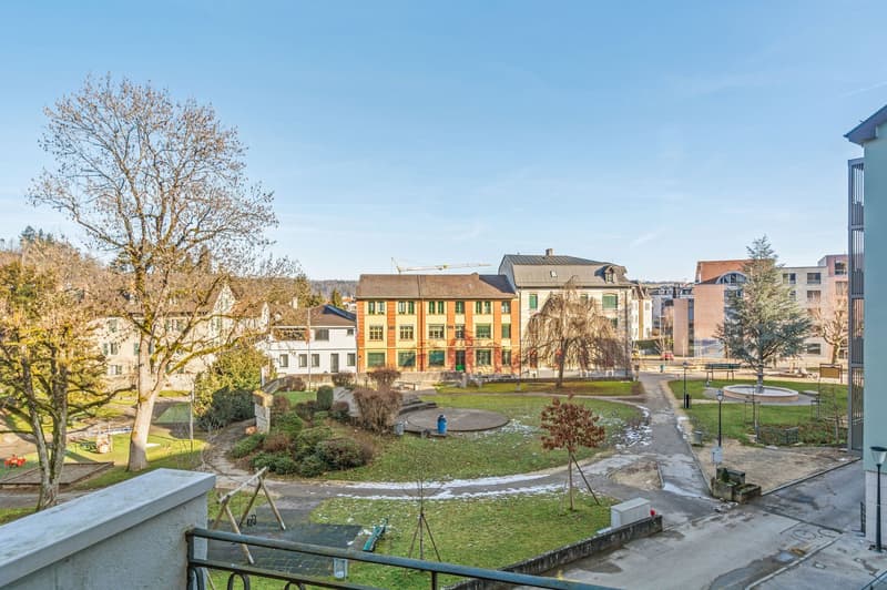 Balcony overlooking a courtyard with grass, trees, benches, and a round structure, with buildings in the background.