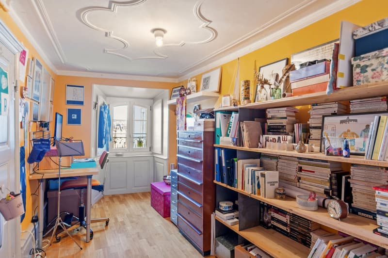 yellow walls, wooden floor, desk, chair, shelves with books, office supplies, framed pictures, window with balcony