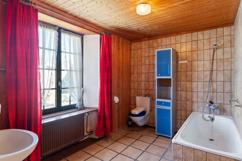Bathroom with wooden walls, tiled floor, bathtub, toilet, sink, window with red curtains, radiator, blue cabinet, shower head