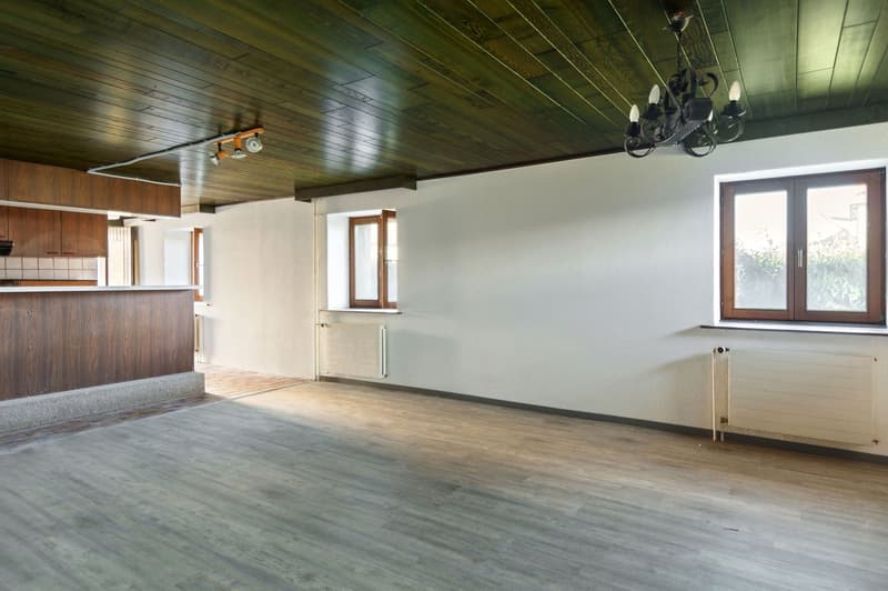 Empty room with wooden floor, white walls, wooden ceiling, two windows, radiators, and a chandelier