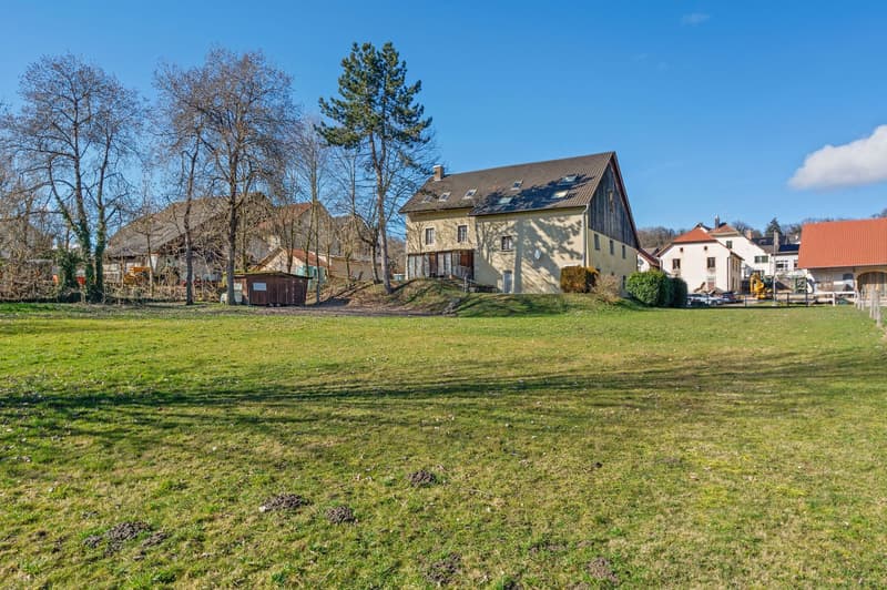 House on a raised ground floor, surrounded by trees and grass, with a large field in front