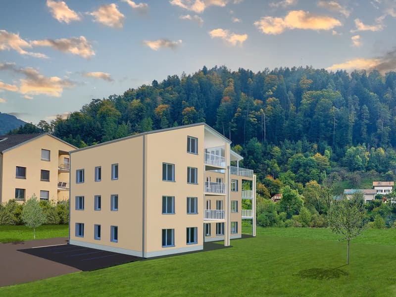 A modern apartment building with beige exterior, multiple windows, and balconies on the upper floors, set in a lush green landscape with a mountain backdrop.
