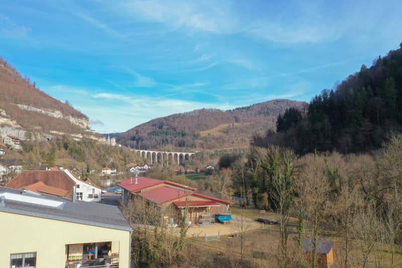 House with red roof, surrounded by trees, mountains, and a river in the background