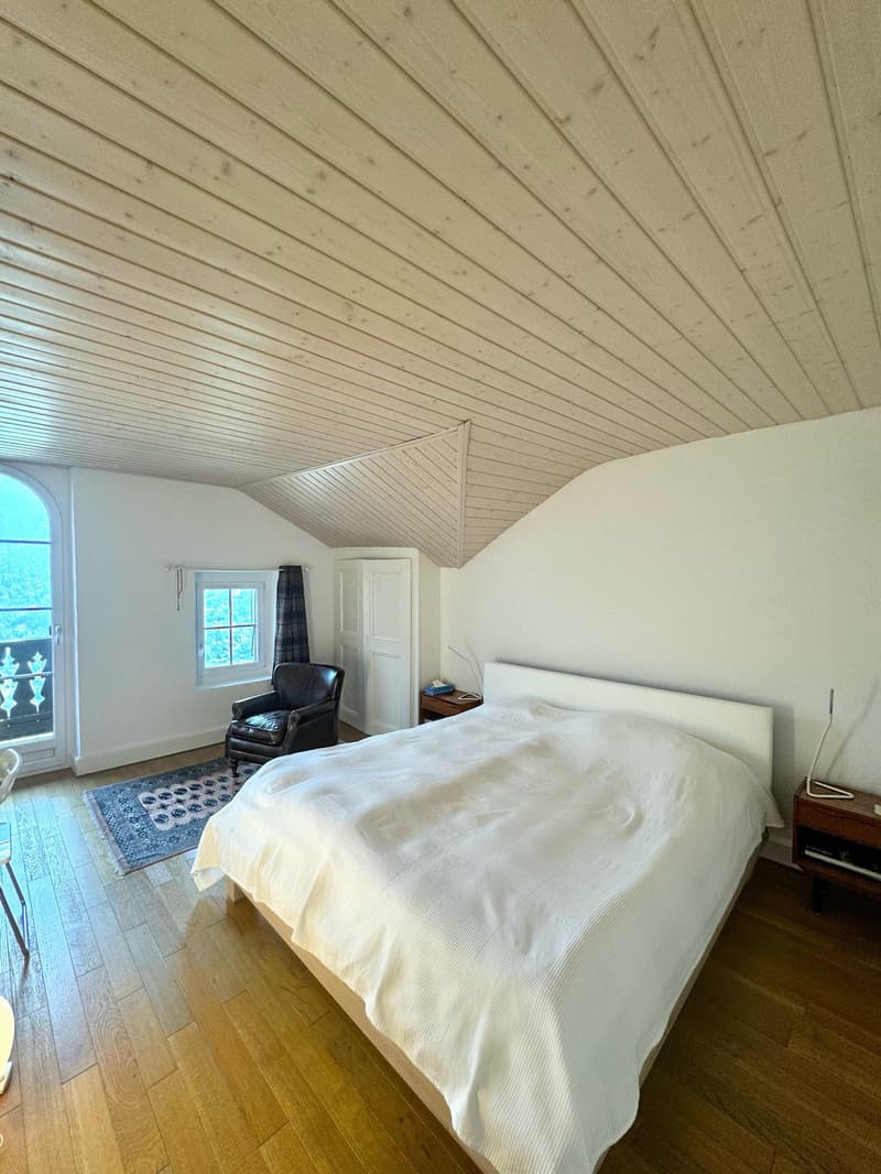 white ceiling, wooden bed, white blanket, wooden floor, leather chair, large window, wooden nightstand, rug, wooden cabinet