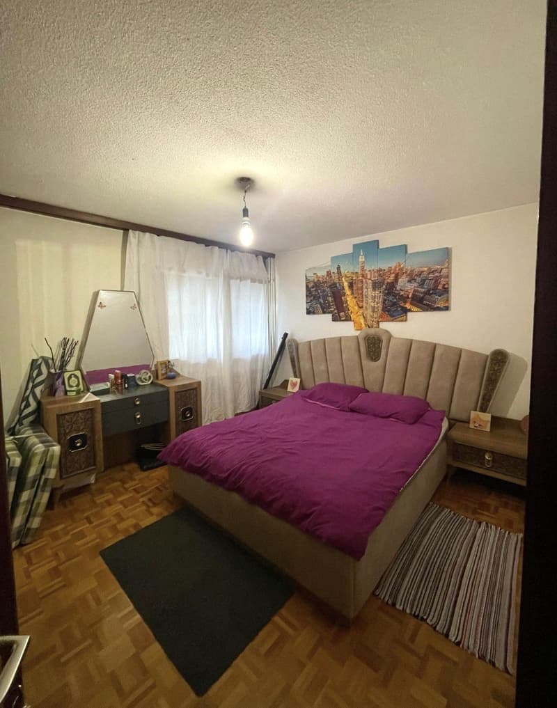 Large bed with purple bedding, wooden dresser, desk, white curtains, rug, city scene painting above bed