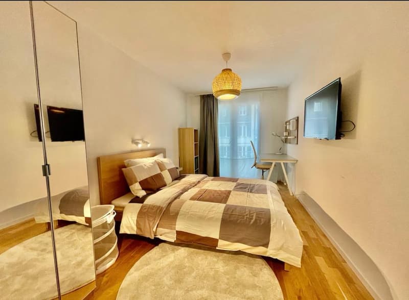 Bedroom with double bed, pillows, blanket, wooden floor, carpet, wall-mounted television, chair, desk, large mirror, lamp, wall-mounted lights, window with curtains.