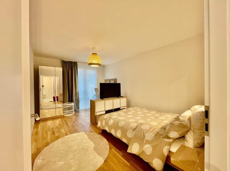 a cozy bedroom with a wooden floor, round rug, bed with a polka dot bedspread, wooden cabinet with a TV, and a mirrored wardrobe, a window with white curtains, and a lamp hanging from the ceiling