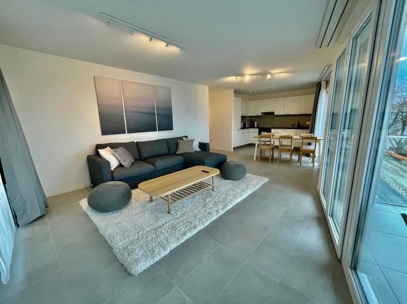 modern living room with large windows, gray couch, wooden table, kitchen