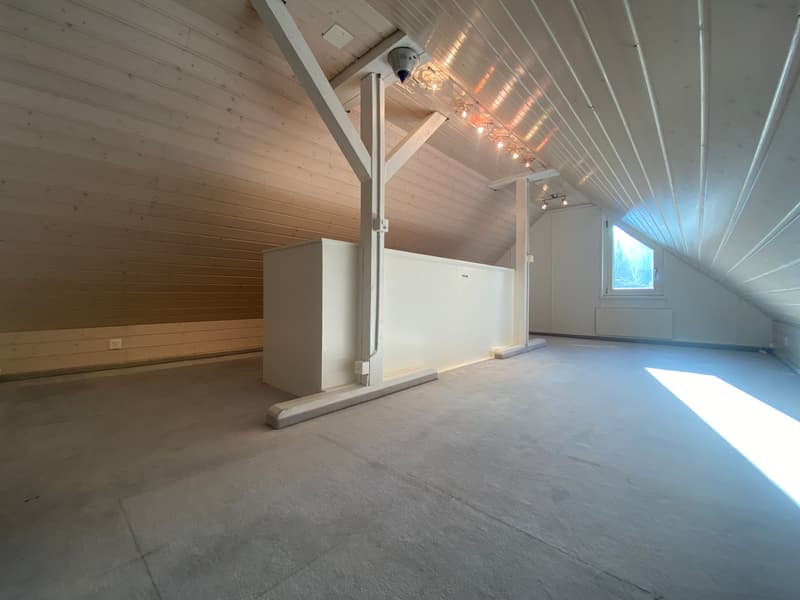 Ample attic room with high ceilings and exposed wooden beams, central heating radiator, ceiling lamps, large window, white-painted walls, gray flooring.