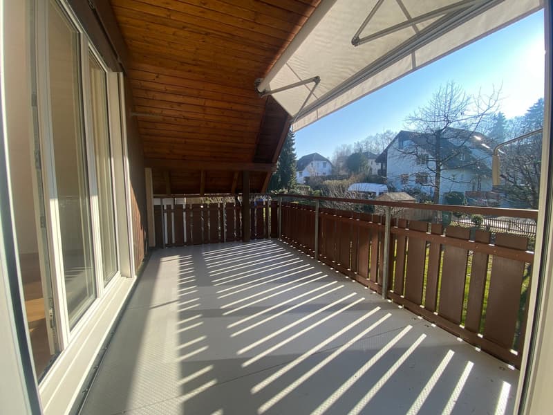 A wooden balcony with a glass railing attached to a house with windows and a wooden ceiling