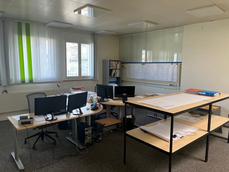 The image shows an office space with multiple desks, computers, and office equipment. The room has large windows, a whiteboard, and shelves with books and documents. The overall layout and furnishings suggest this is a professional work environment.