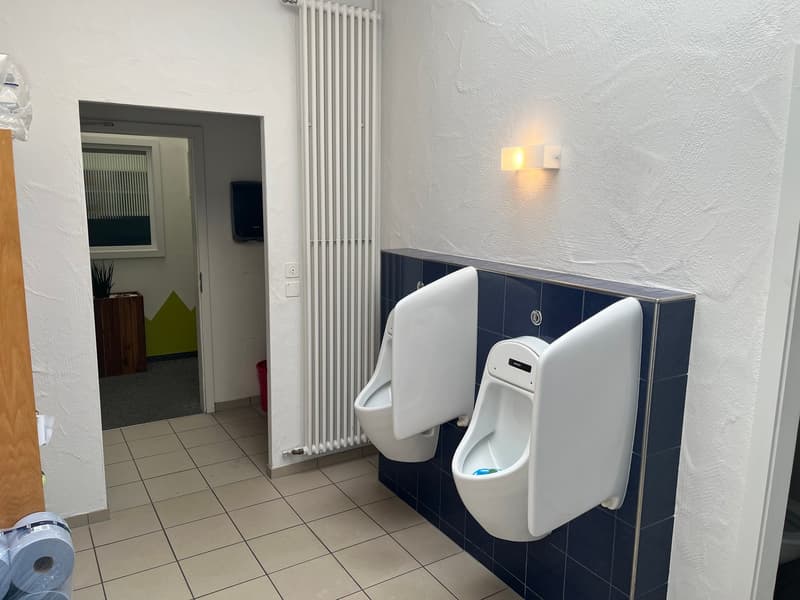 Tiled floor, white urinals, radiator, doorway leading to another room