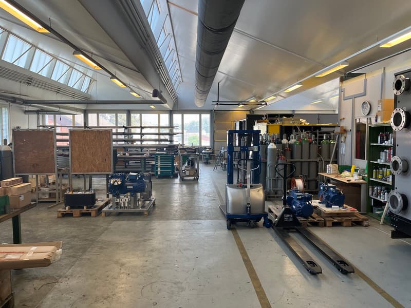 Large industrial workshop space with high ceilings, concrete floors, and various tools and equipment such as shelves, workbenches, and machinery. The space appears to be used for manufacturing or repair work.