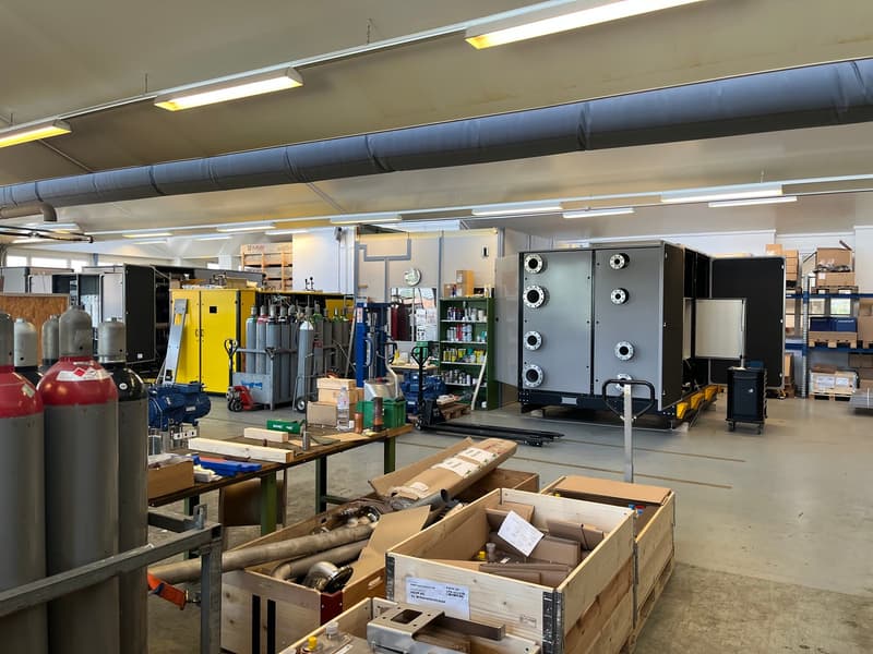 The image shows a large industrial warehouse or storage facility. The space is filled with various equipment, tools, and supplies, including large metal tanks, shelves, and boxes. The ceiling has exposed ductwork and lighting fixtures. There appears to be