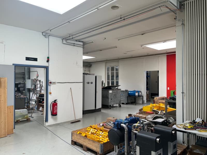 The image shows a large, open garage or workshop space with various tools, equipment, and storage containers visible. The space has a high ceiling with overhead lighting and storage racks. There is a large garage door that appears to be open, allowing acc