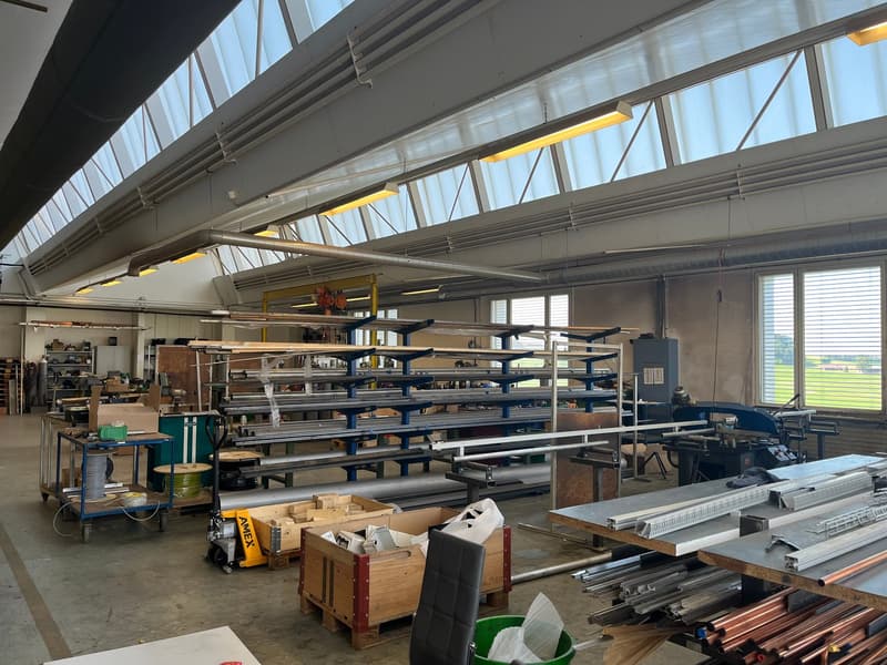 Large industrial workshop space with high ceilings, shelving units, workbenches, and various tools and equipment. The space appears to be used for storage and manufacturing, with a garage door visible indicating it may be part of a larger industrial or co