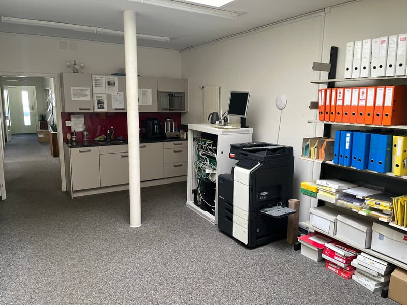 The image shows an office space with a kitchen area, a printer/copier machine, and various storage shelves and cabinets. The kitchen area has a red backsplash and appears to have a dishwasher. The office space has a carpeted floor and contains various off