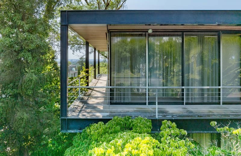 Modern design, large windows, metal railing, wooden floor, elevated position, greenery, and view of the surrounding area