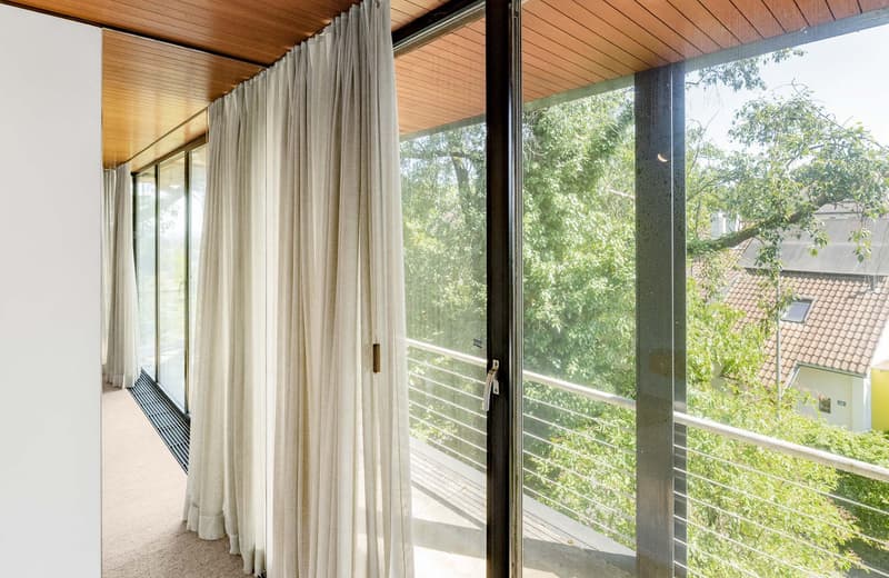 Balcony with glass doors and metal railings, beige flooring, sliding doors, beige curtain, trees outside