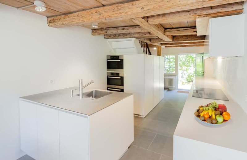Modern kitchen with white cabinets, stainless steel appliances including a built-in oven, and a sink with a faucet. The kitchen has exposed wooden beams on the ceiling, creating a rustic feel. There is a bowl of fresh fruits on the counter.