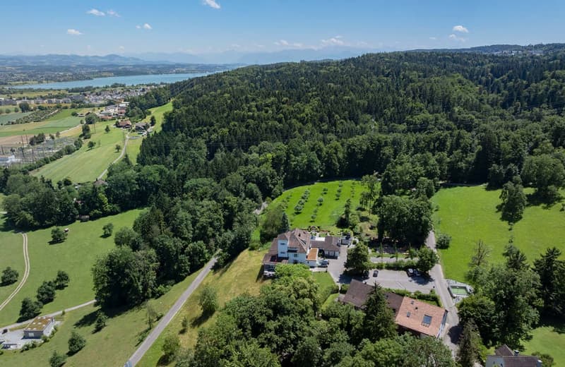 Aerial view of a scenic countryside landscape with a lake, rolling hills, forests, and a residential area with houses and buildings nestled among the greenery.